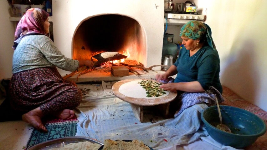 Two women in traditional Turkish clothing prepare food at home, one sitting near a wood-fired hearth and the other kneading dough on a low table with various utensils around.