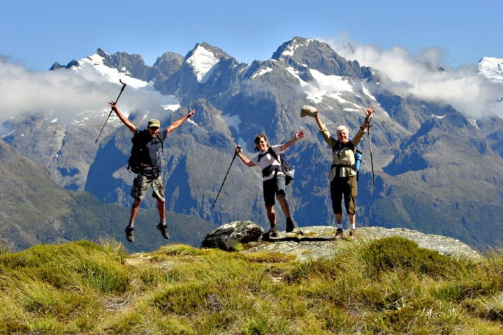Three hikers with trekking poles stand on a rocky outcrop in New Zealand, two jumping with arms raised, with a breathtaking mountainous landscape in the background.