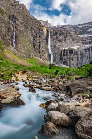 A scenic view of a waterfall cascading down a rocky cliff into a stream surrounded by rocks and green grass, with a blue sky and clouds in the background, perfect for tourism and travel enthusiasts exploring the charming landscapes of France.