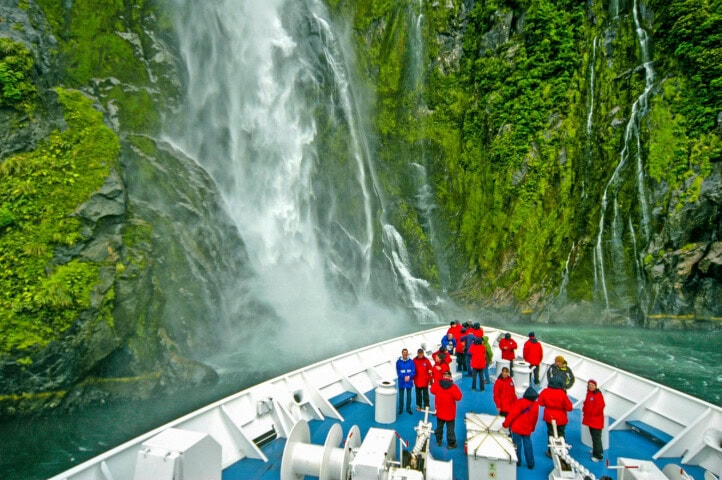 A group of people in red jackets stand on a boat deck approaching a large waterfall cascading down a green, moss-covered cliff in New Zealand.