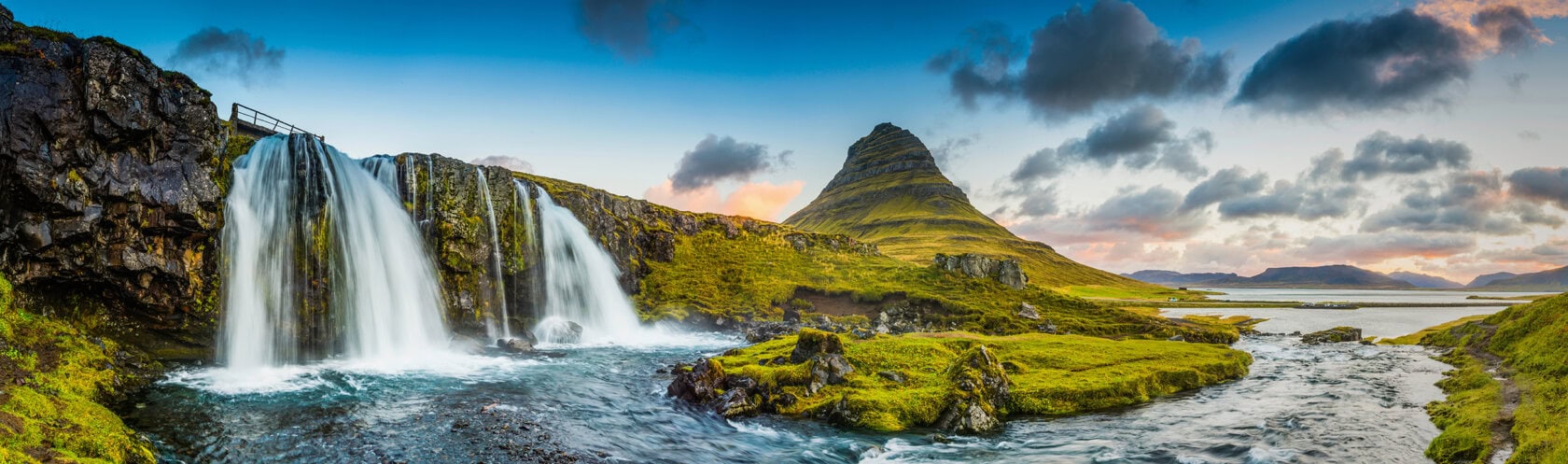 Panoramic view of a waterfall flowing over rocks with a grassy landscape, river, and a conical mountain under a partly cloudy sky.