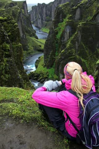 A person with blond hair in a pink jacket and backpack sits on a grassy ledge, overlooking a moss-covered canyon with a winding river, capturing the essence of Iceland's natural beauty—a true paradise for travel and tourism.