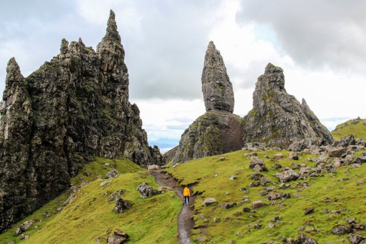 A person in a yellow jacket walks along a narrow path through jagged rock formations and green grassy terrain under a cloudy sky, capturing the essence of Scotland's rugged beauty—a perfect scene for travel and tourism enthusiasts.