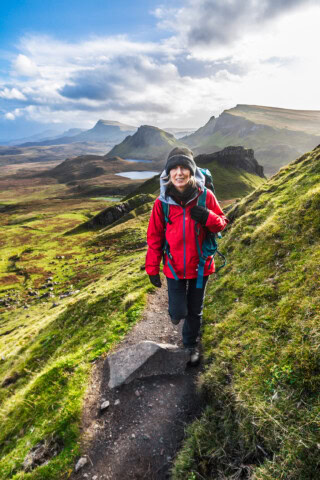 A hiker in a red jacket and backpack treks along a narrow trail through the vast, green mountainous landscape of Scotland, with a serene lake and cloudy sky in the background.