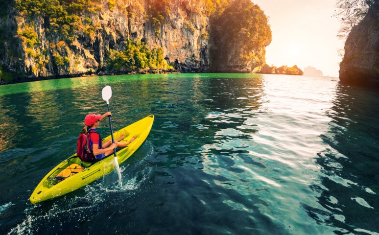 Person kayaking on calm water in Palau, surrounded by tall cliffs and greenery, with the sun setting in the background—an idyllic travel destination that captures the essence of nature's beauty.