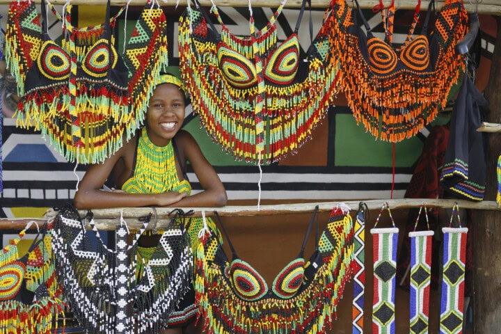 A traveler smiling behind a wooden stall displays colorful beaded ornaments and accessories, sharing tales from their latest trip.