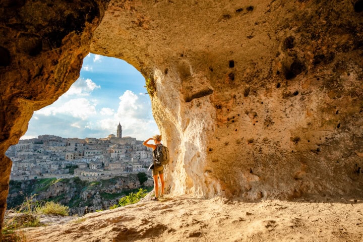 A person stands in a rocky cave, looking out through an opening at an ancient city on a hillside under Italy's blue sky with scattered clouds.