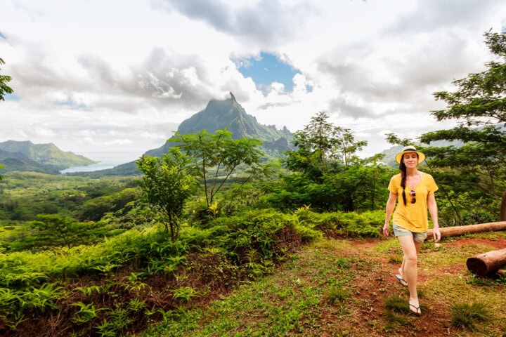 A woman in a yellow shirt and hat walks through the lush green landscape of French Polynesia, with mountains and a cloudy sky in the background.