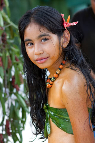 A girl with long dark hair adorned with a flower and painted in traditional attire, including a leaf top and beaded necklace, looks at the camera, capturing the essence of Palau's rich cultural heritage.