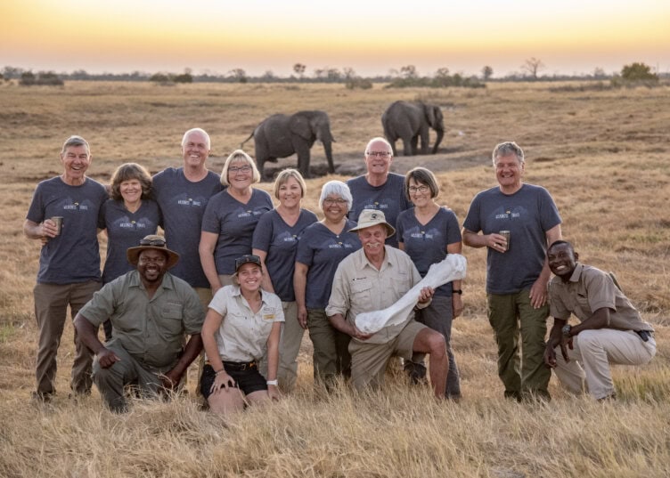 A group of travelers and safari guides pose for a photo in a grassy field at sunset, with elephants grazing peacefully in the background.